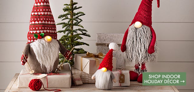 An image of three fabric gnomes with red hats and glowing noses on a table with gifts. Shop Indoor Holiday Decorations